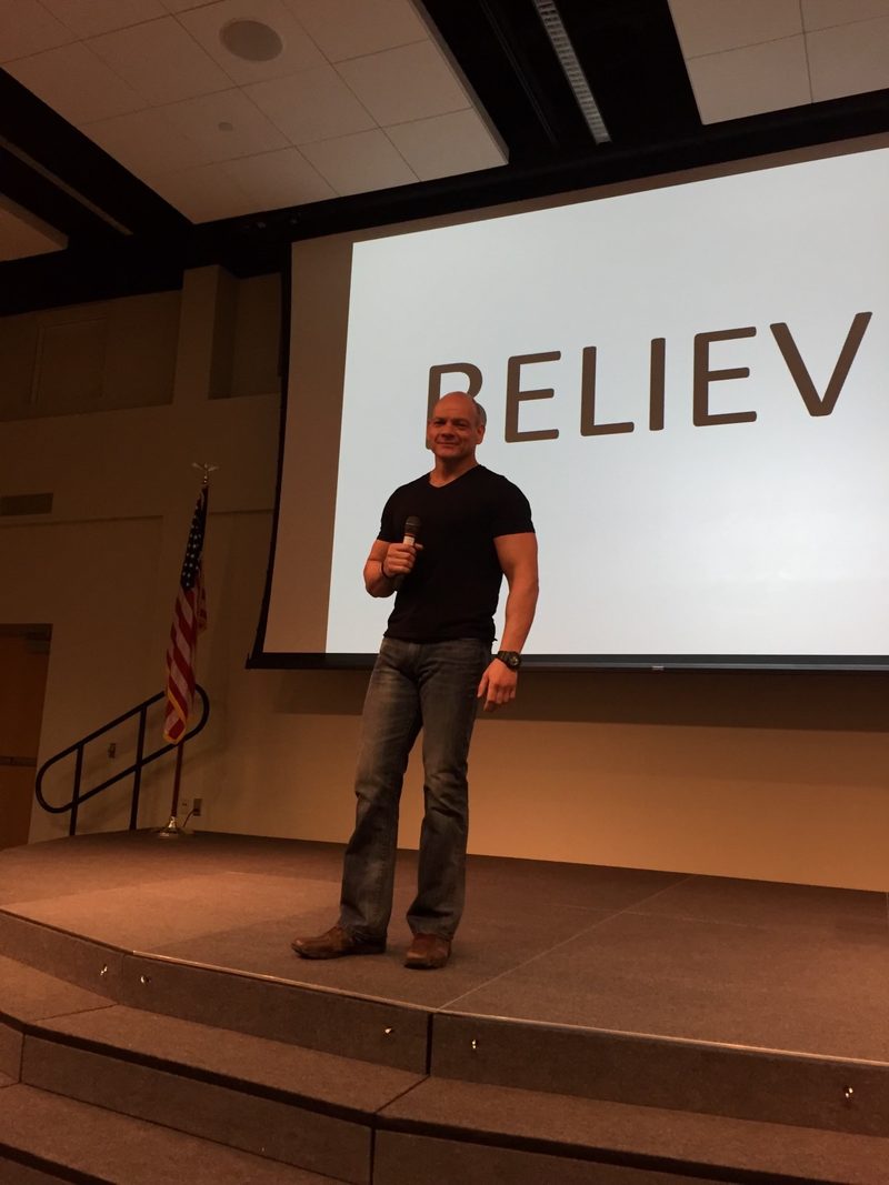 Phil speaking on a stage in front of a screen that reads BELIEVE